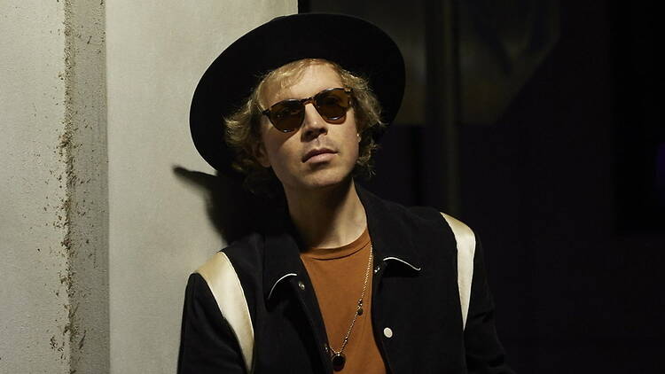 A close-up of a person wearing a hat and sunglasses against a dark backdrop.