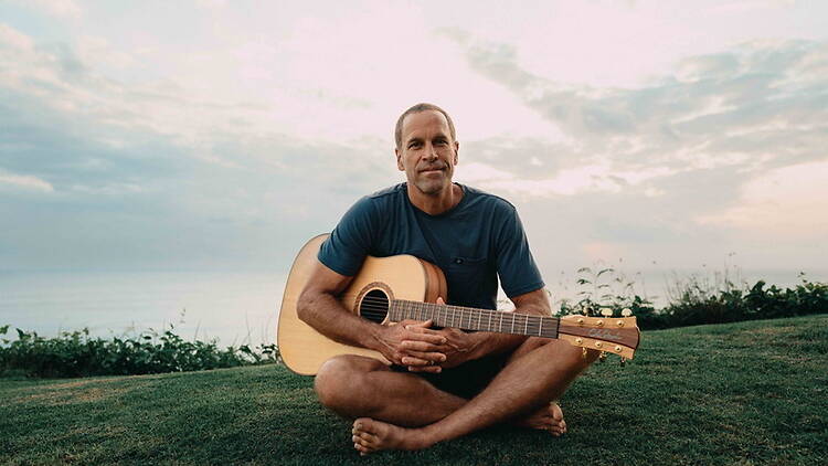 A wide shot of a person sitting with an acoustic guitar on grass by the water.