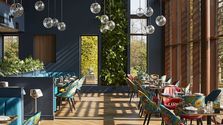 the main dining room on the property, featuring teal banquette seating, botanical floral-patterned chairs, globe pendant lights, and a living green wall.