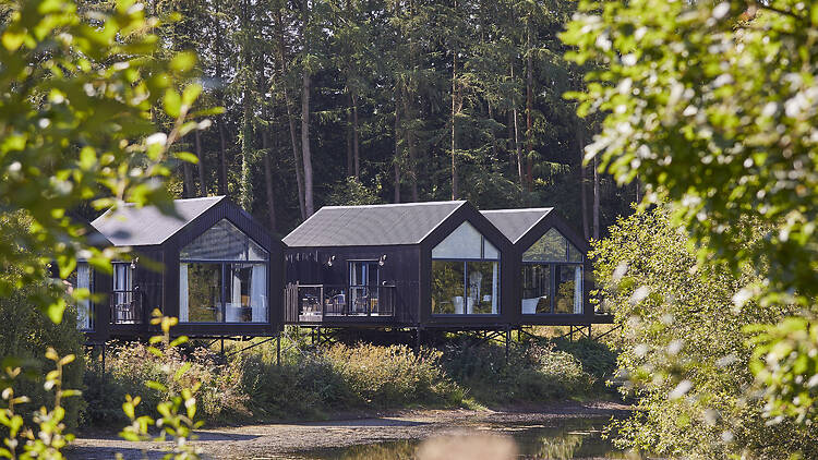standalone, black timber-clad cabins raised on stilts along the edge of the lake, featuring floor-to-ceiling glass windows and private outdoor decking with outdoor spa baths