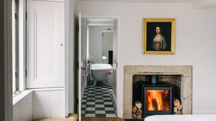 An elegant hotel bedroom featuring a lit wood-burning stove set inside a stone fireplace, with a framed portrait hanging above. An open doorway leads into an en-suite bathroom with black-and-white checkered floor tiles and a freestanding clawfoot bathtub.