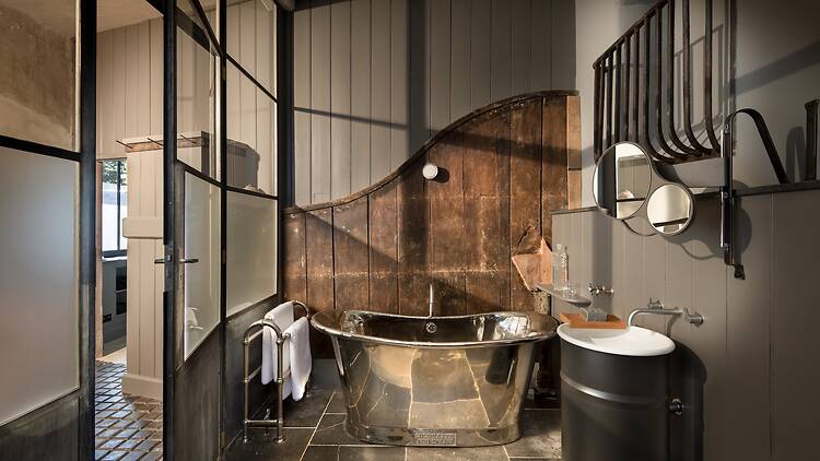 A rustic luxury bathroom featuring a polished metallic bathtub set against a curved wooden panel and grey-painted wall. Black metal-framed glass doors frame the entrance, alongside a pedestal sink crafted from a dark drum, topped with a circular mirror.