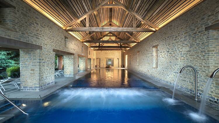 An indoor-outdoor swimming pool set within a rustic stone barn structure with vaulted timber ceiling beams. Sunlight and water streams flow through the pool area, which leads out toward an exterior terrace lined with lounge chairs.
