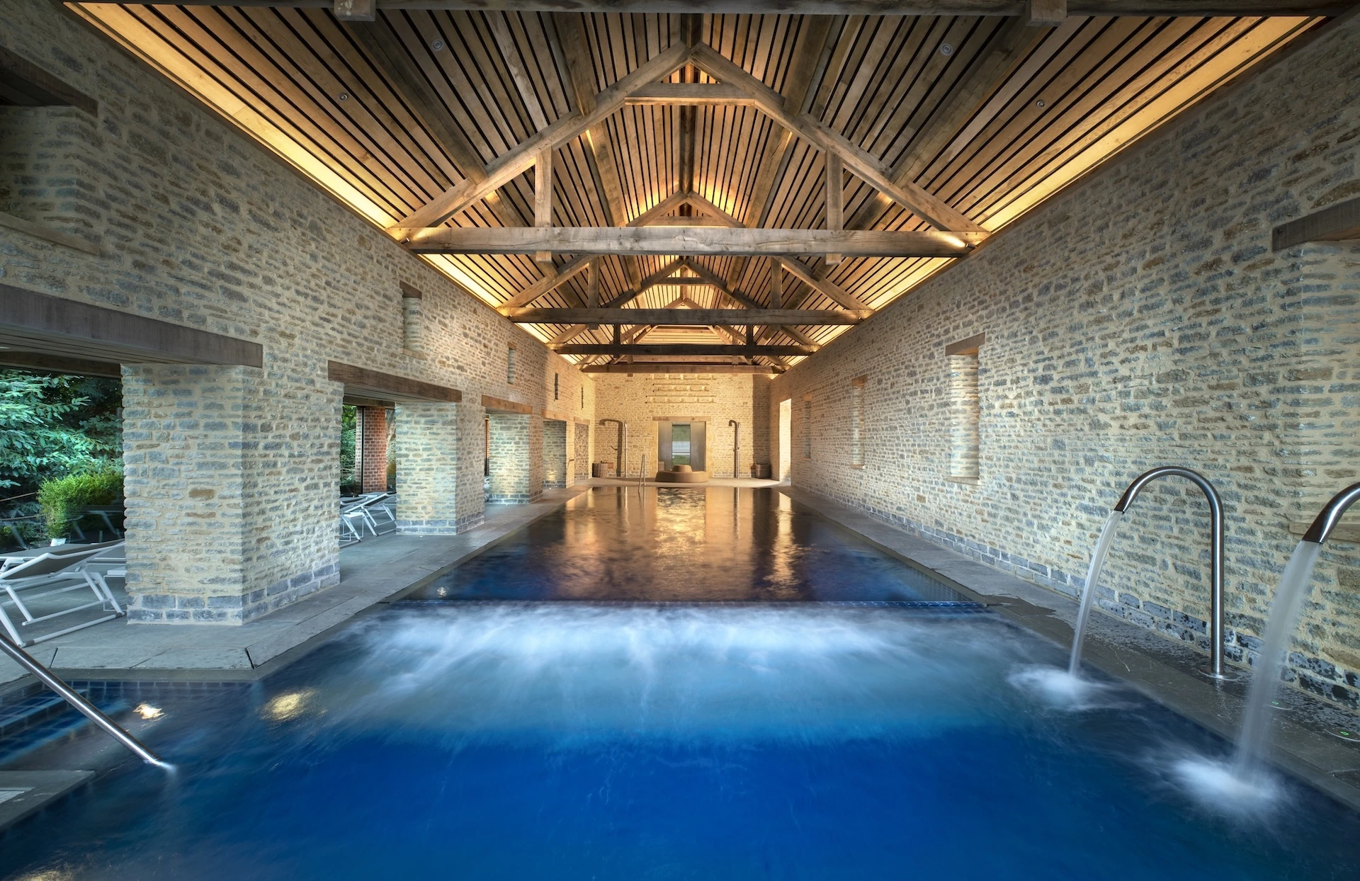 An indoor-outdoor swimming pool set within a rustic stone barn structure with vaulted timber ceiling beams. Sunlight and water streams flow through the pool area, which leads out toward an exterior terrace lined with lounge chairs.