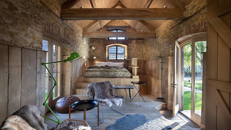 A cozy rustic-luxury hotel room inside a converted stone barn with vaulted timber ceilings and exposed brick arches. Steps lead up to an elevated bed area, while the lower seating area