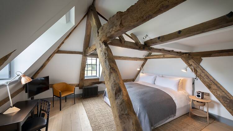 An attic-style hotel bedroom featuring exposed, original wooden timber beams framing the bed and sloped ceilings