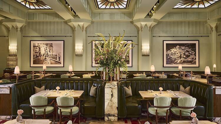 An elegant Art Deco-style restaurant dining room featuring deep-green leather booths, mint-green velvet armchairs, marble tables, and illuminated glass ceiling panels above pale green walls.
