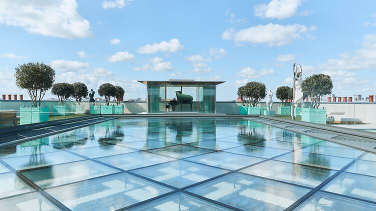A modern rooftop terrace with a large glass skylight floor over water, potted trees, and a grand piano housed inside a central glass pavilion against a clear blue sky.