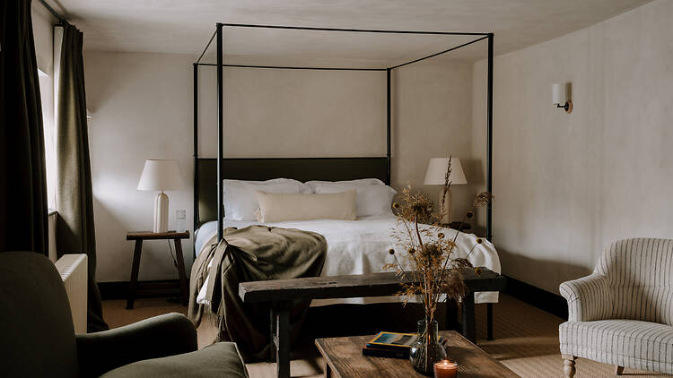 A cozy, warm-toned bedroom in a rustic countryside pub featuring a black metal four-poster bed made up with white linens