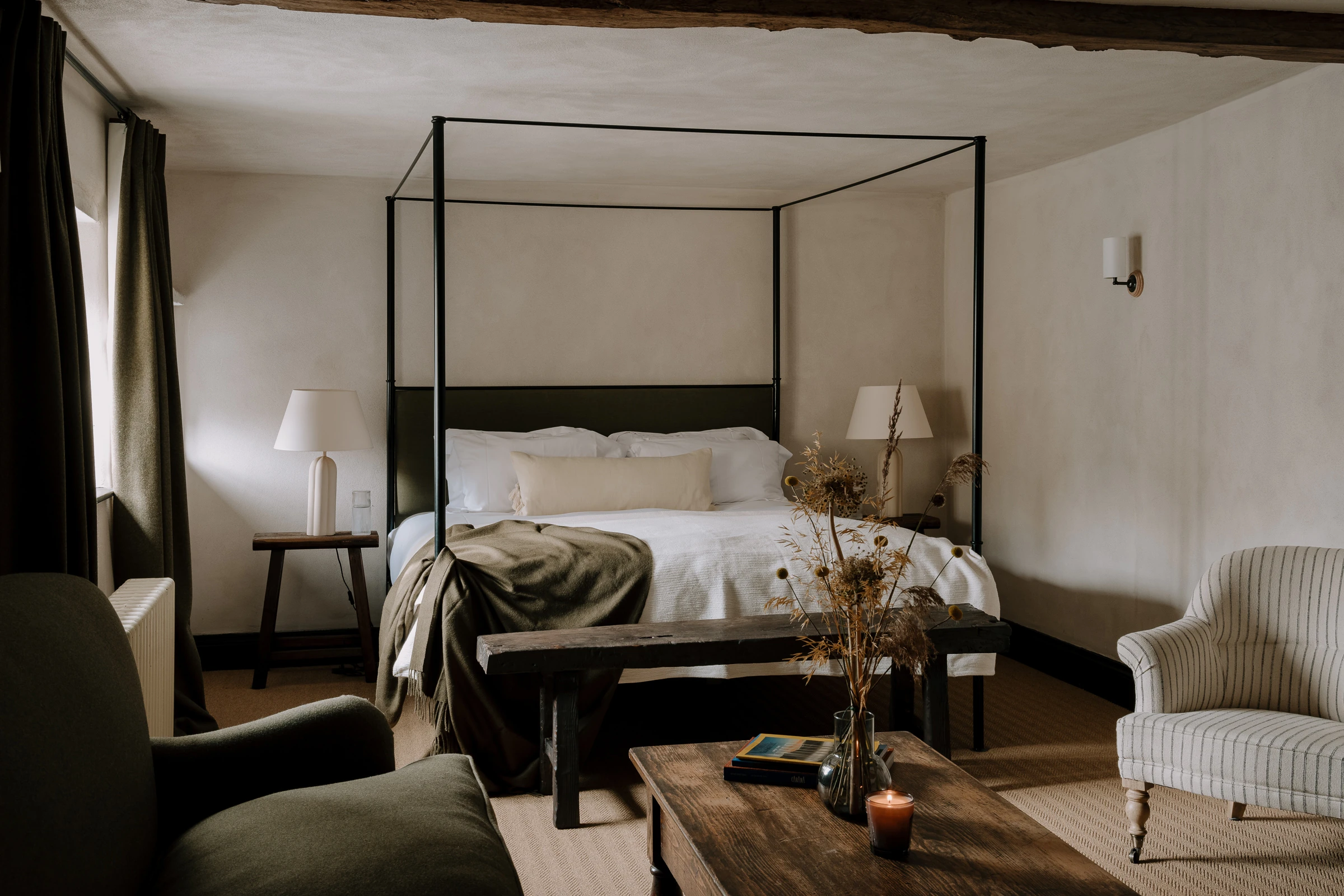 A cozy, warm-toned bedroom in a rustic countryside pub featuring a black metal four-poster bed made up with white linens