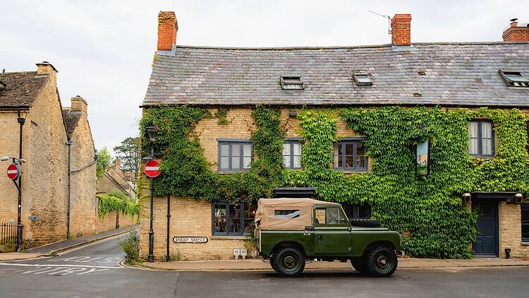 The exterior of a historic Cotswold stone building covered in lush green ivy with grey-framed glass windows and a tiled slate roof