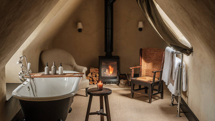A cozy attic bathroom featuring sloping plaster walls, a black clawfoot bathtub with a wooden bath tray, and a burning wood-burning stove in the center background
