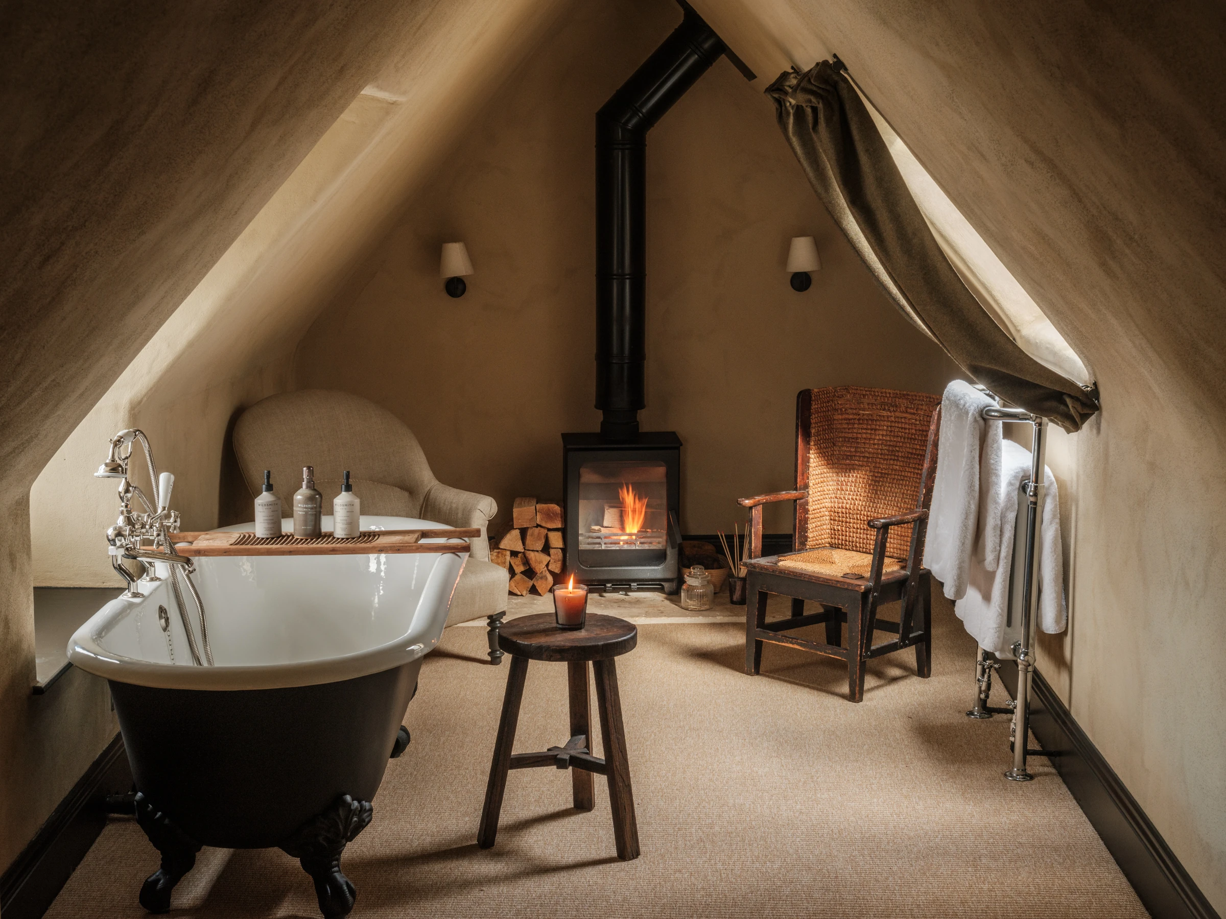 A cozy attic bathroom featuring sloping plaster walls, a black clawfoot bathtub with a wooden bath tray, and a burning wood-burning stove in the center background