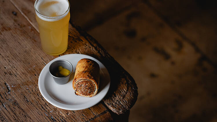 A white plate of a sausage roll and mustard next to a half pint