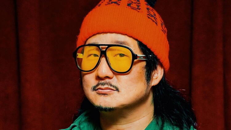 A close-up of a person wearing an orange beanie and yellow-tinted glasses against a dark red backdrop.