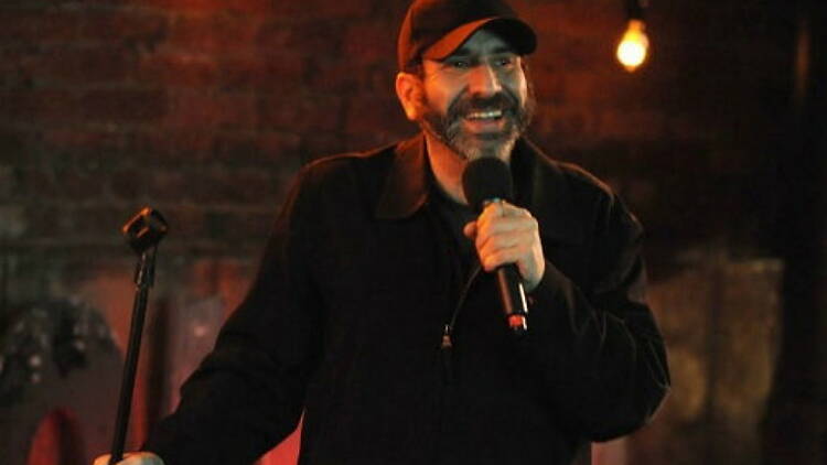 A close-up of a comedian smiling and speaking into a microphone on stage. A close-up of a comedian smiling and speaking into a microphone on stage.
