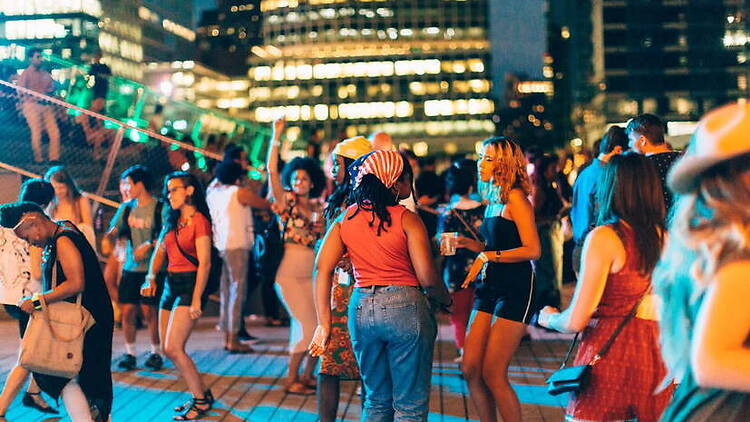 A wide shot of people dancing and socializing at an outdoor nighttime gathering in a city plaza A wide shot of people dancing and socializing at an outdoor nighttime gathering in a city plaza