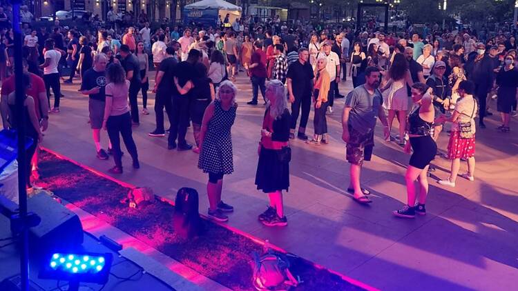 A wide shot of people socializing and dancing at an outdoor evening gathering in a city plaza