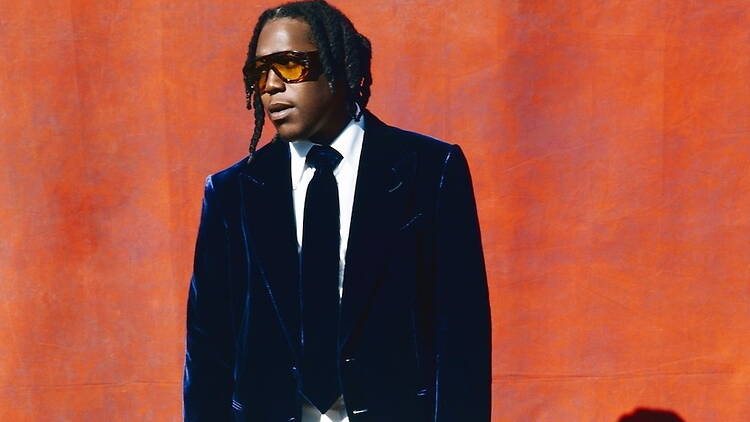 Close-up of a person standing against an orange backdrop while wearing a suit and sunglasses
