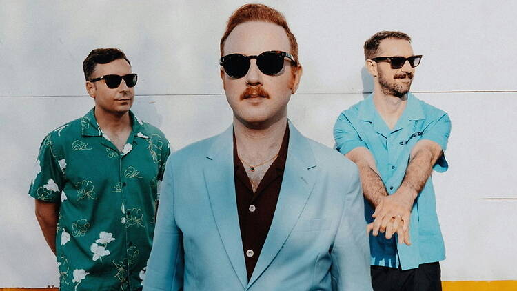 A wide shot of three people posing outdoors against a light-colored wall while wearing sunglasses A wide shot of three people posing outdoors against a light-colored wall while wearing sunglasses