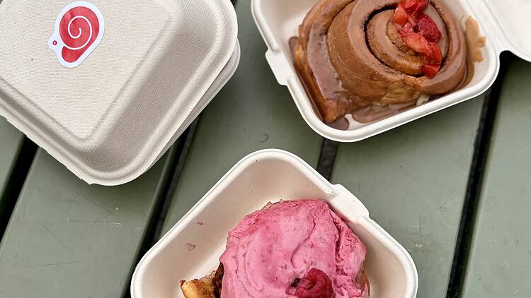 Fresh Buns from viral Bunzy Cafe in Stockholm Takeout containers on a green table featuring fresh American-style cinnamon and sticky buns from Bunzy in Stockholm, including one topped with pink raspberry frosting and another with glaze and strawberries.