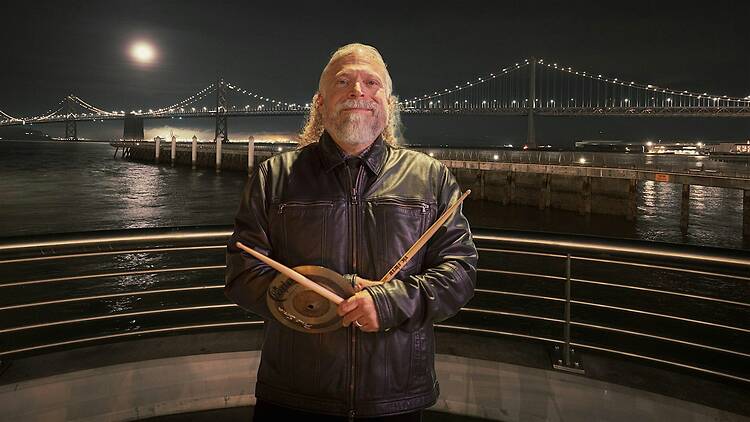 A wide shot of a person holding a pair of drumstick and a small cymbal by the waterfront at night. A wide shot of a person holding a pair of drumstick and a small cymbal by the waterfront at night.