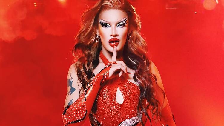 A close-up of a performer posing in a red sequined outfit under red stage lights. A close-up of a performer posing in a red sequined outfit under red stage lights.