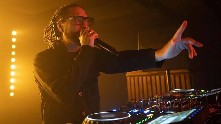A wide shot of a DJ speaking into a microphone while performing at a mixing console. A wide shot of a DJ speaking into a microphone while performing at a mixing console.