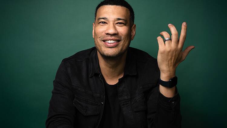 A close-up of a person smiling and raising a hand against a green backdrop. A close-up of a person smiling and raising a hand against a green backdrop.