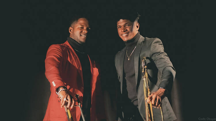A wide shot of two musicians posing with saxophones against a dark backdrop.