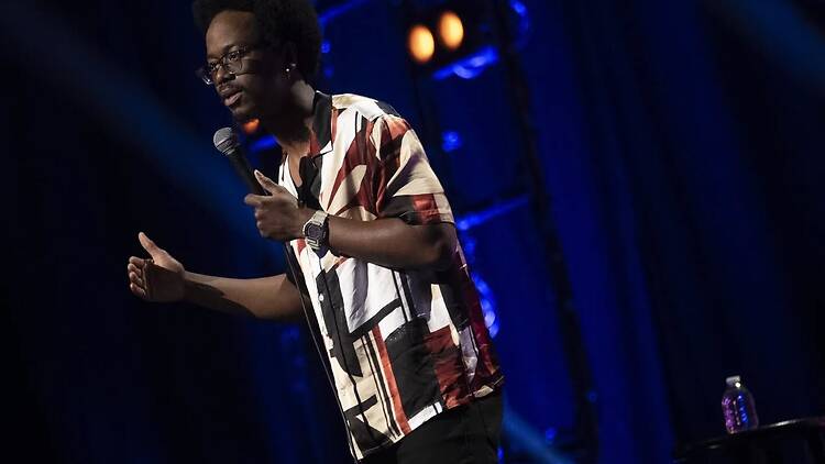A wide shot of a comedian speaking into a microphone and gesturing on stage. A wide shot of a comedian speaking into a microphone and gesturing on stage.