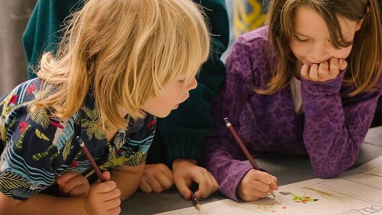 A close-up of children drawing together on a large sheet of paper at a table. A close-up of children drawing together on a large sheet of paper at a table.