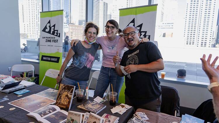 A wide shot of people posing behind a display table at a zine festival in a convention hall A wide shot of people posing behind a display table at a zine festival in a convention hall
