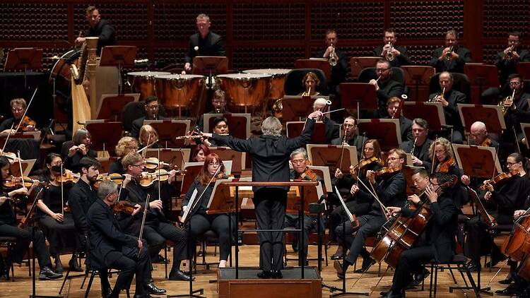 A wide shot of musicians performing under a conductor’s direction on a concert hall stage A wide shot of musicians performing under a conductor’s direction on a concert hall stage