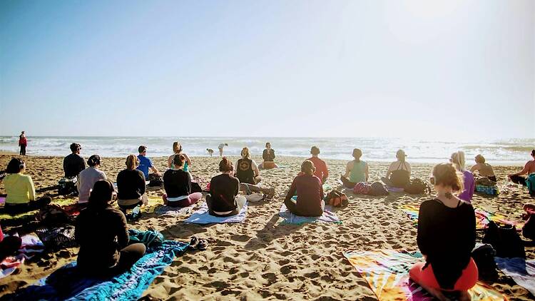 A wide shot of people meditating together on a beach overlooking the ocean.