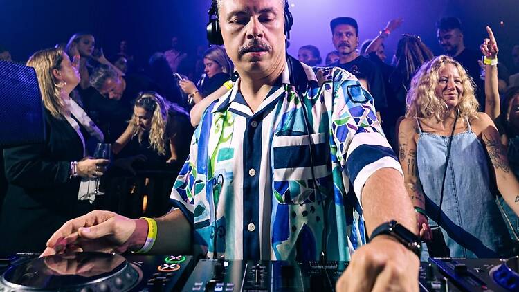 A wide shot of a DJ mixing music at a crowded indoor event. A wide shot of a DJ mixing music at a crowded indoor event.