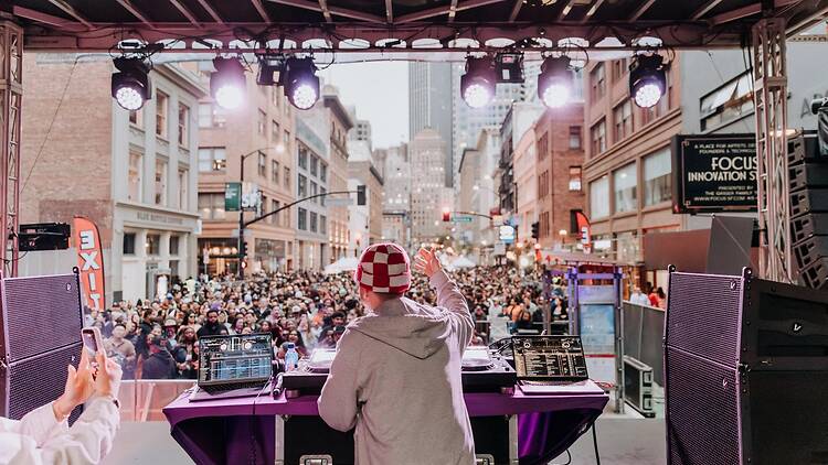 A wide shot of a DJ performing on an outdoor stage for a large crowd in the city.