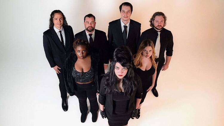 A wide shot of six people posing together in formal black outfits against a light backdrop. A wide shot of six people posing together in formal black outfits against a light backdrop.