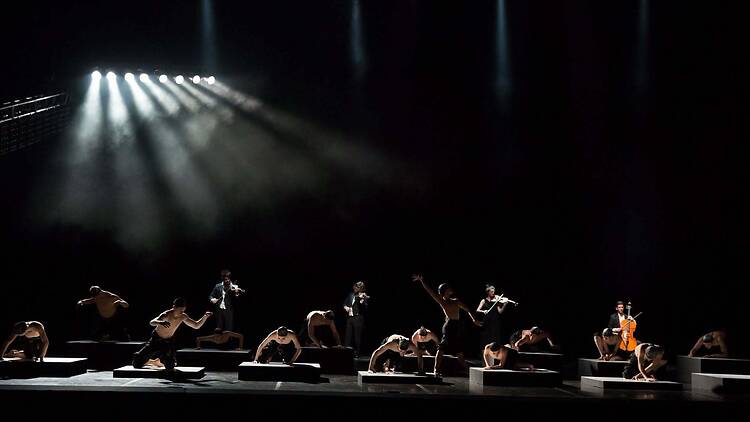 A wide shot of dancers performing with violinists on a dark theater stage. A wide shot of dancers performing with violinists on a dark theater stage.