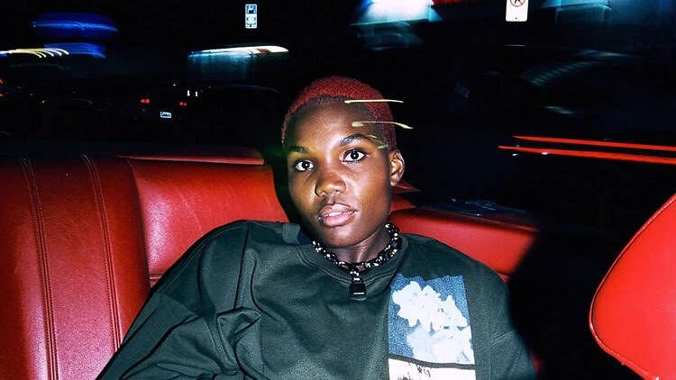 A close-up of a person sitting in a car, wearing a dark shirt and necklace.