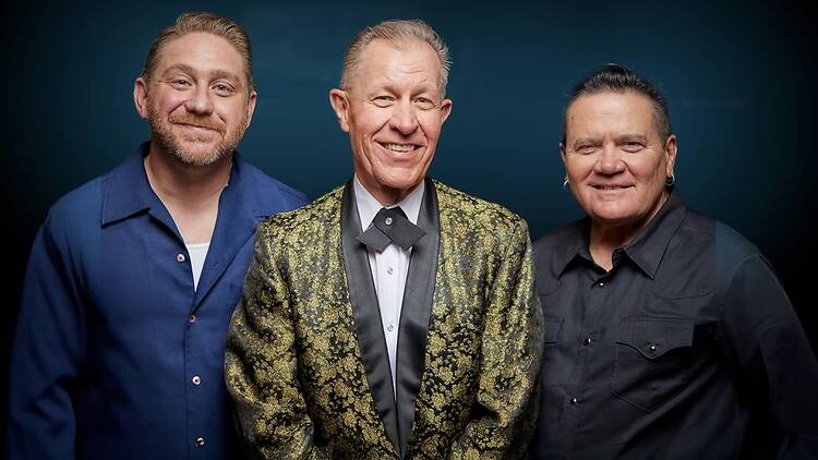 A close-up of three people posing together in front of a dark blue backdrop. A close-up of three people posing together in front of a dark blue backdrop.