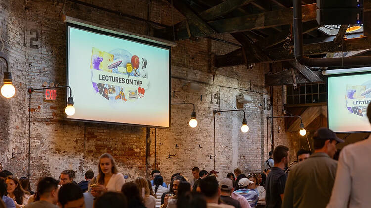 A wide shot of a crowd attending a lecture event inside a brick-walled venue. A wide shot of a crowd attending a lecture event inside a brick-walled venue.