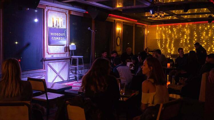 A wide shot of an audience seated in a comedy club facing a stage. A wide shot of an audience seated in a comedy club facing a stage.