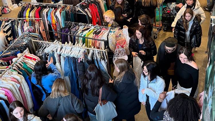 A wide shot of shoppers browsing clothing racks at an indoor clothing market. A wide shot of shoppers browsing clothing racks at an indoor clothing market.