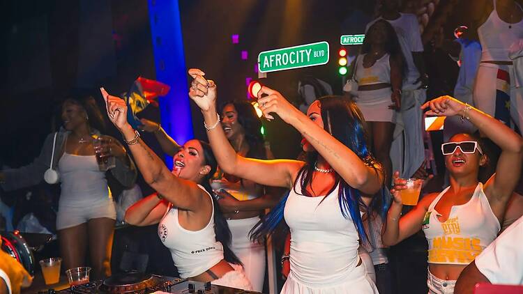 A wide shot of people dancing and cheering around a DJ booth at an indoor event. A wide shot of people dancing and cheering around a DJ booth at an indoor event.