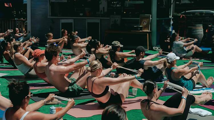 A wide shot of people practicing a seated stretching exercise together outdoors. A wide shot of people practicing a seated stretching exercise together outdoors.