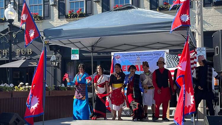 A wide shot of people in traditional Nepali attire performing on an outdoor stage with Nepali flags. A wide shot of people in traditional Nepali attire performing on an outdoor stage with Nepali flags.