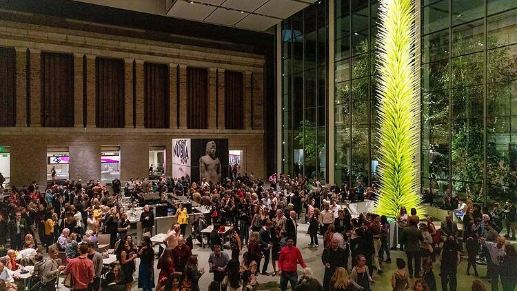 A wide shot of a crowd socializing beneath a large illuminated art installation at an indoor event.