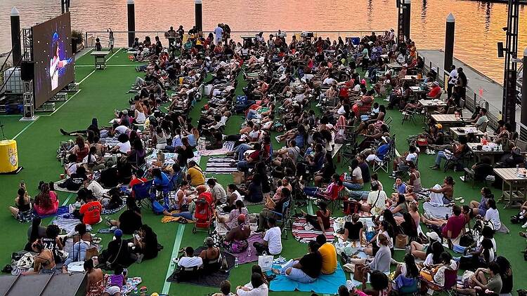 A wide shot of a large crowd watching an outdoor movie screening by the waterfront. A wide shot of a large crowd watching an outdoor movie screening by the waterfront.