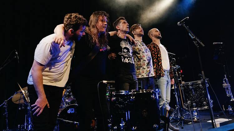 A wide shot of five musicians standing together on stage after a performance.